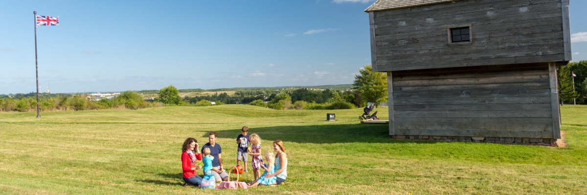 At a distance, a family sit on a blanket picnicking on the lawn near the wooden blockhouse.